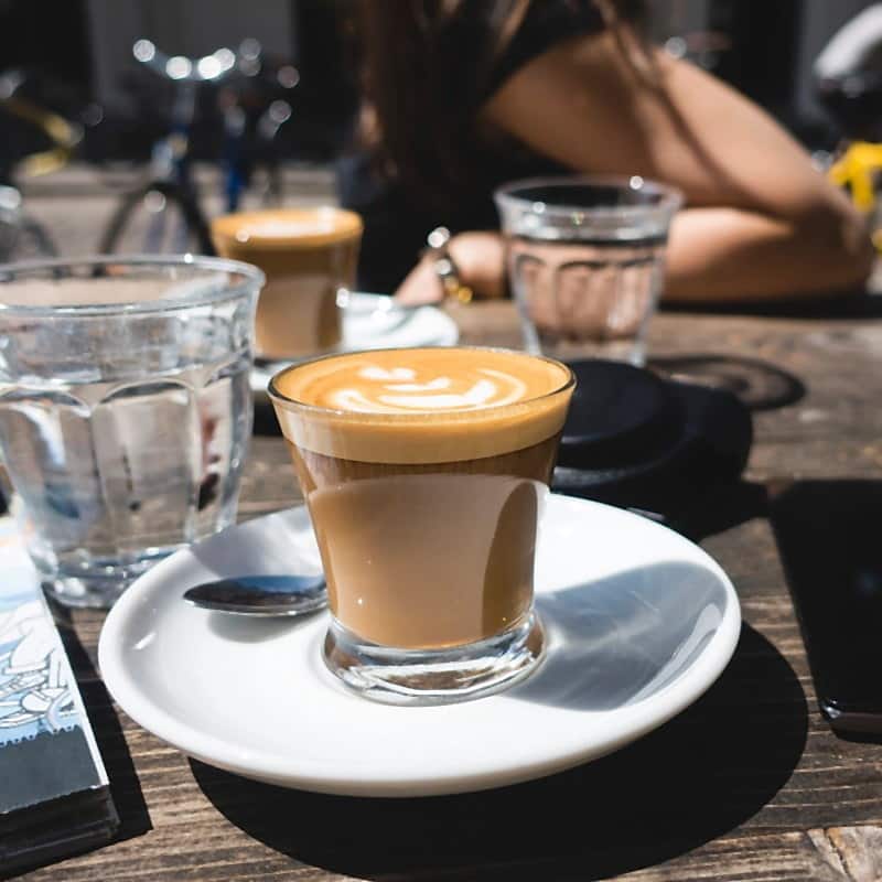 A flat white in a glass tumbler on a saucer, on a café table