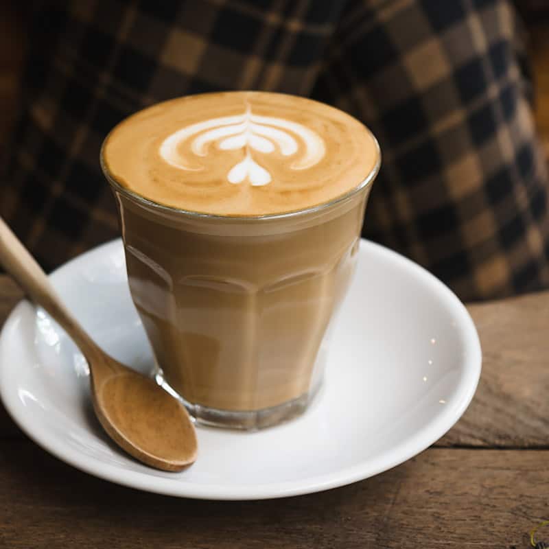 A latte in a fluted glass on a white saucer beside a wooden spoon